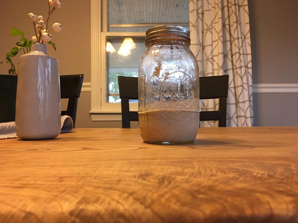 A narrow mouth mason jar sits on a wood table with its metal lid attached. The outside lighting is dim so the dining room light is turned on to show the starter inside the jar.