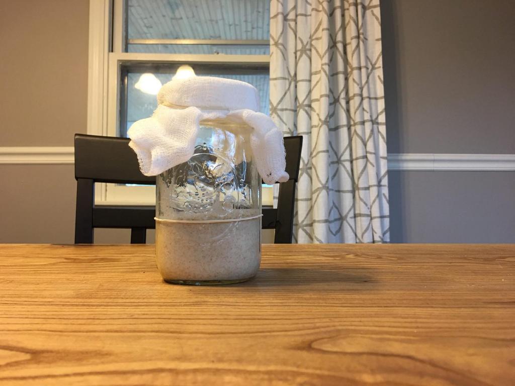 A wide mouth mason jar covered with a cheese cloth is sitting on a raw edge wooden table. The sourdough starter inside doesn't show much activity as it hasn't shown any signs of rising past the rubber band marker